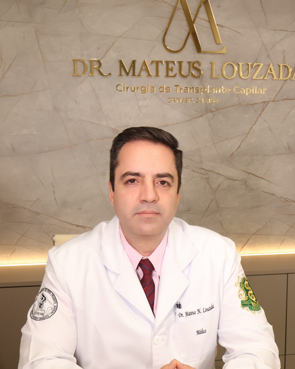 Dr. Mateus Louzada sentado à mesa do consultório, com o letreiro da marca ao fundo